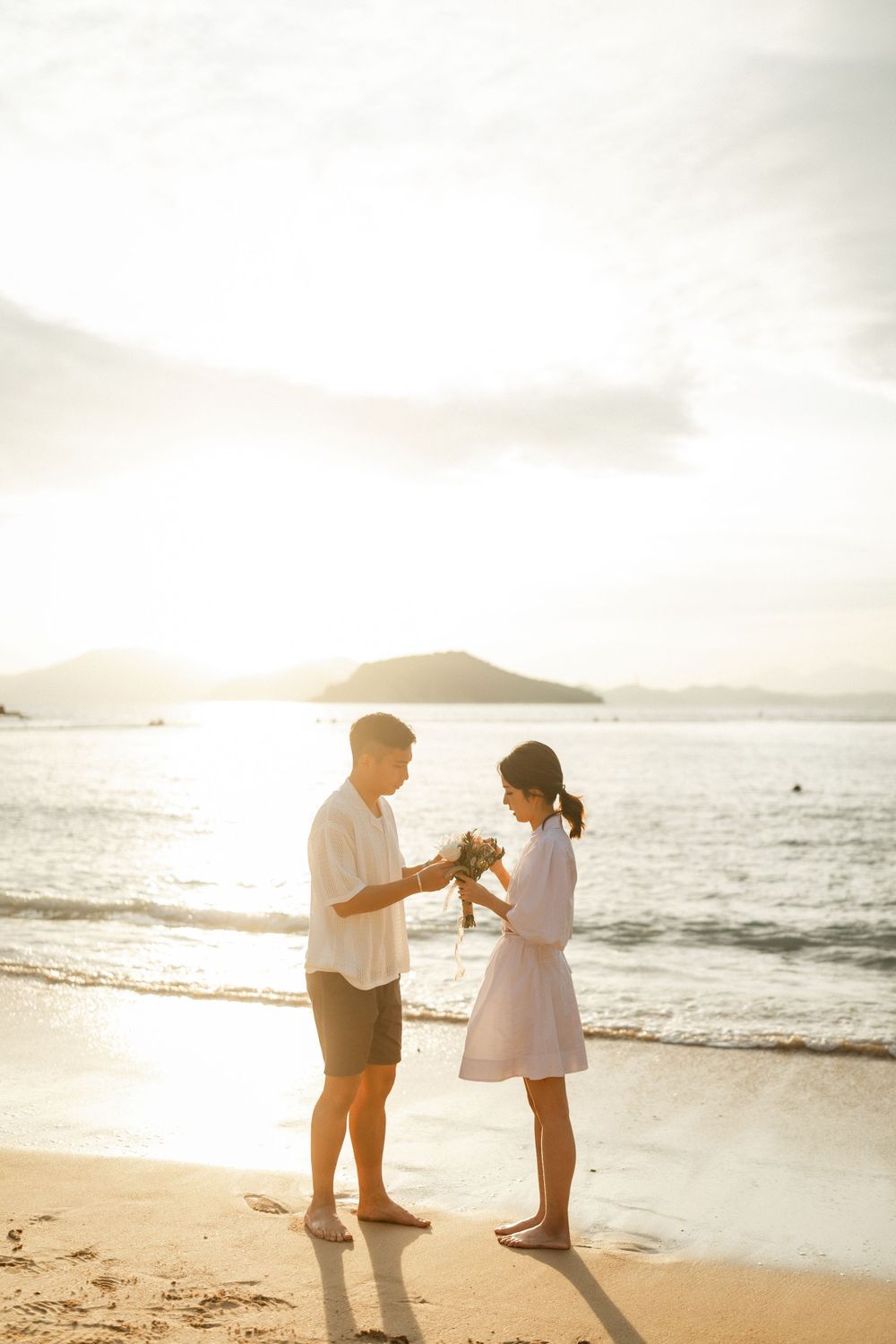 Romantic couple exchanging flowers on beach at sunset with glowing ocean and mountains behind.