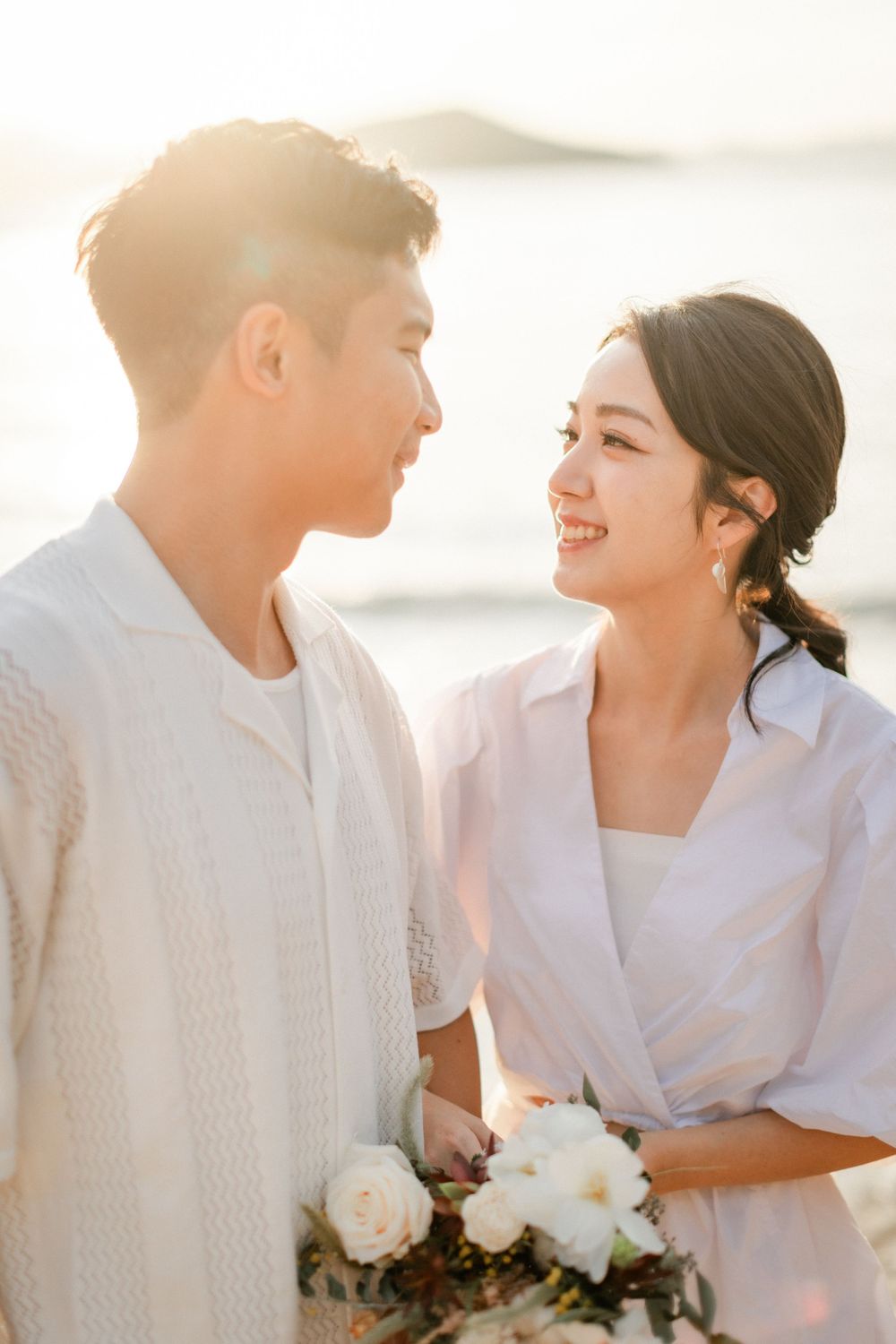 Loving Asian couple gazing at each other holding white roses on beach at golden sunset.