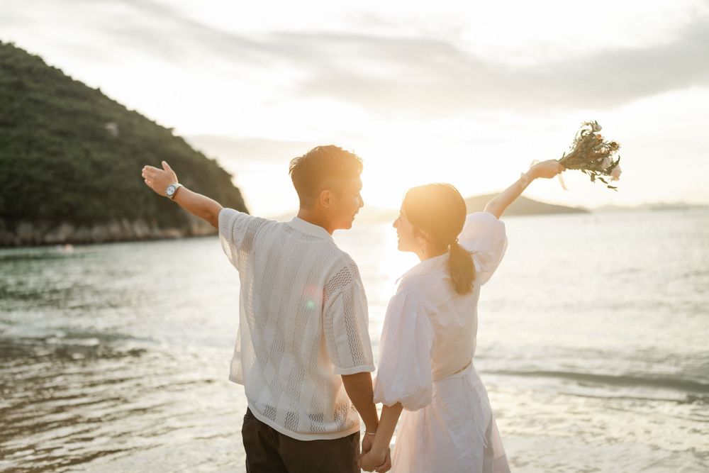 Happy couple holding hands with arms raised holding bouquet on beach at glowing sunset.