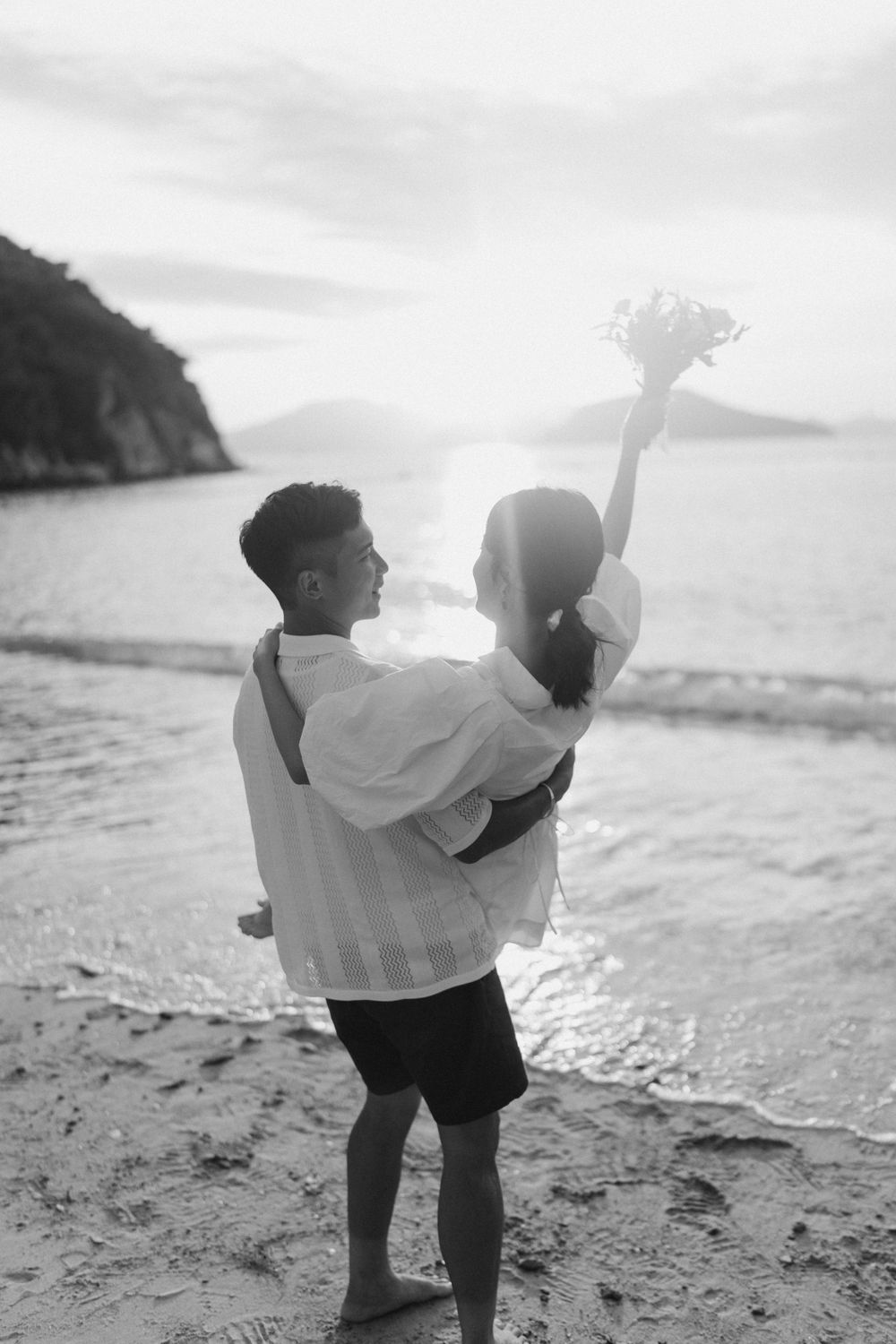 Black and white photo of couple embracing on beach at sunset, woman raising bouquet high.