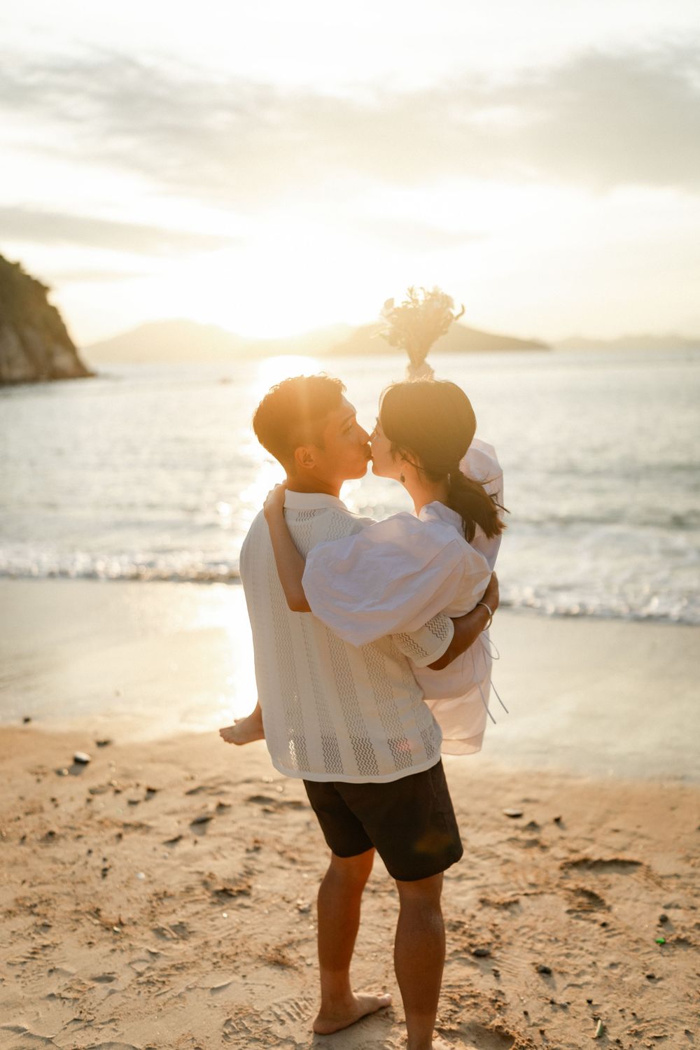Romantic couple kissing while man lifts woman holding bouquet on sandy beach at sunset.