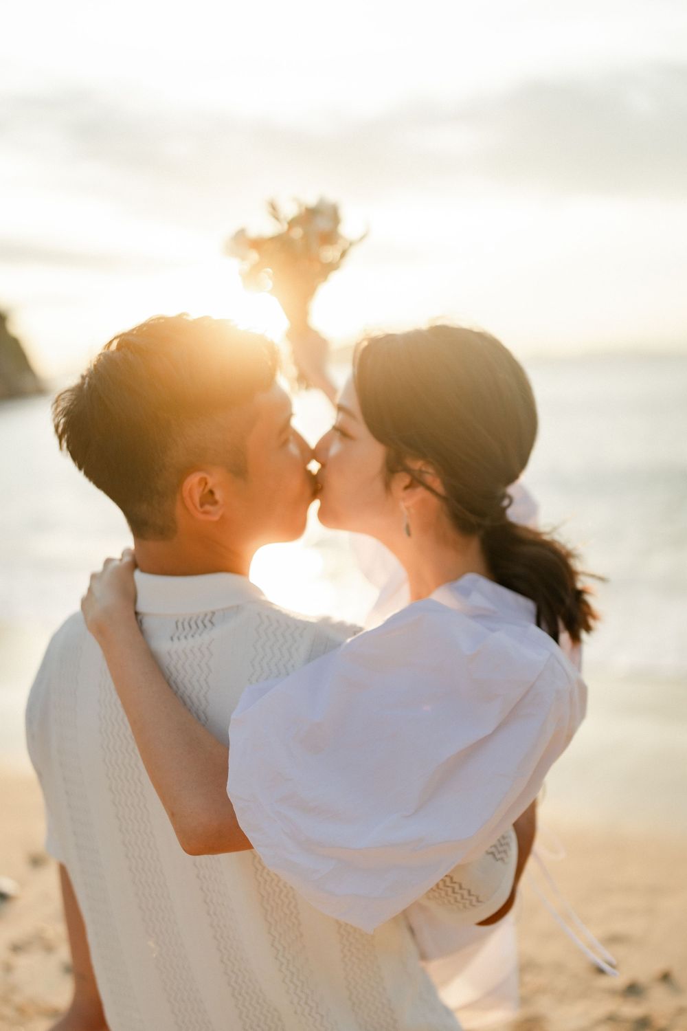 Couple sharing a romantic kiss on the beach at golden sunset, woman holding a bouquet of flowers.