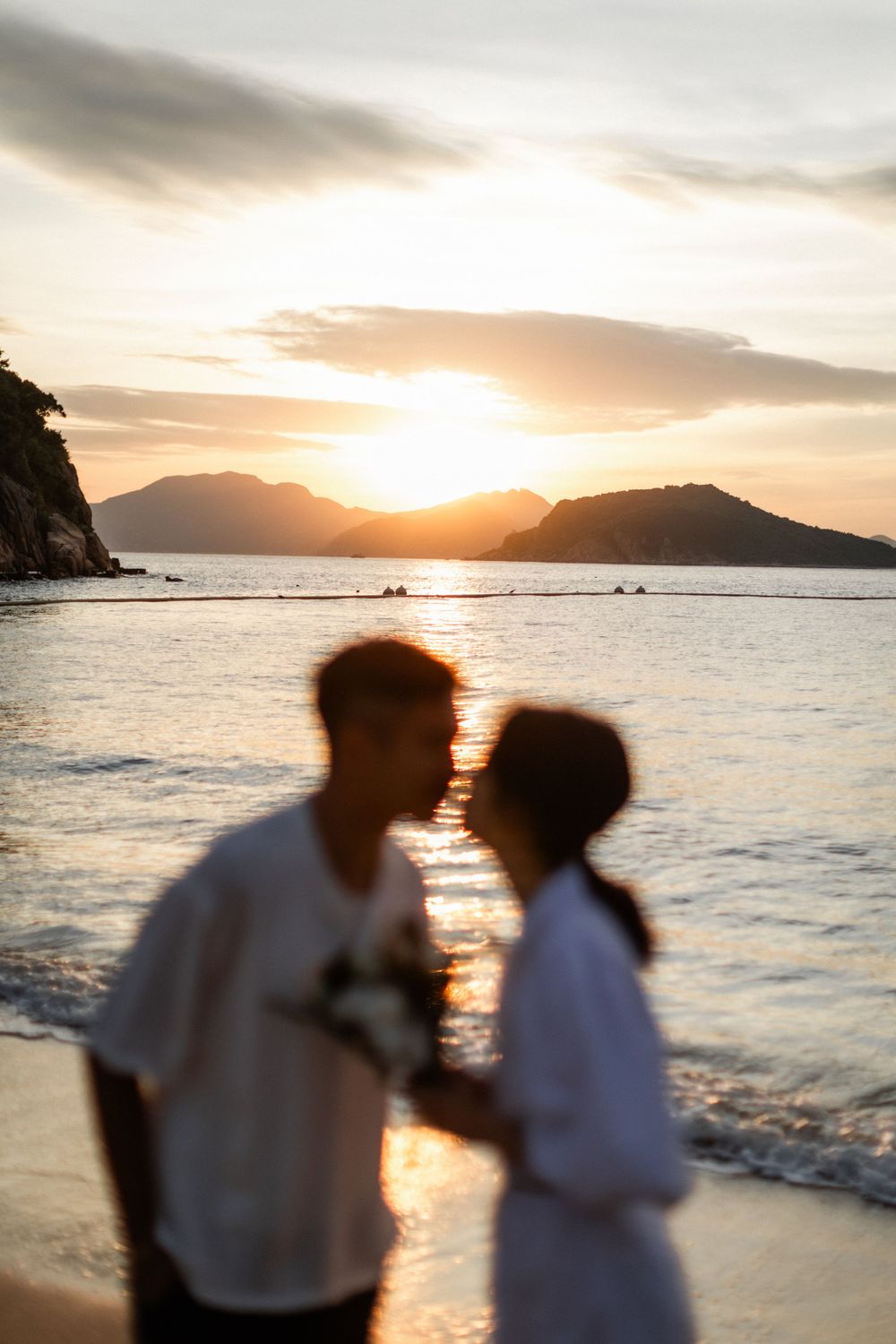 Couple standing close together on a beach at sunset with mountains and ocean in the background.