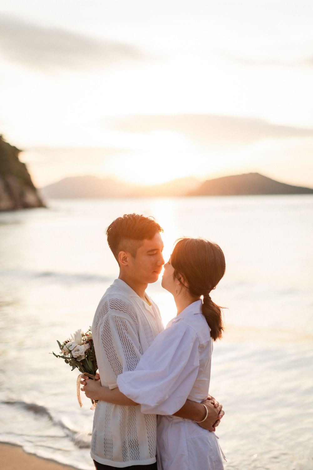 Romantic couple embracing and kissing on a sandy beach during a warm golden sunset by the sea.