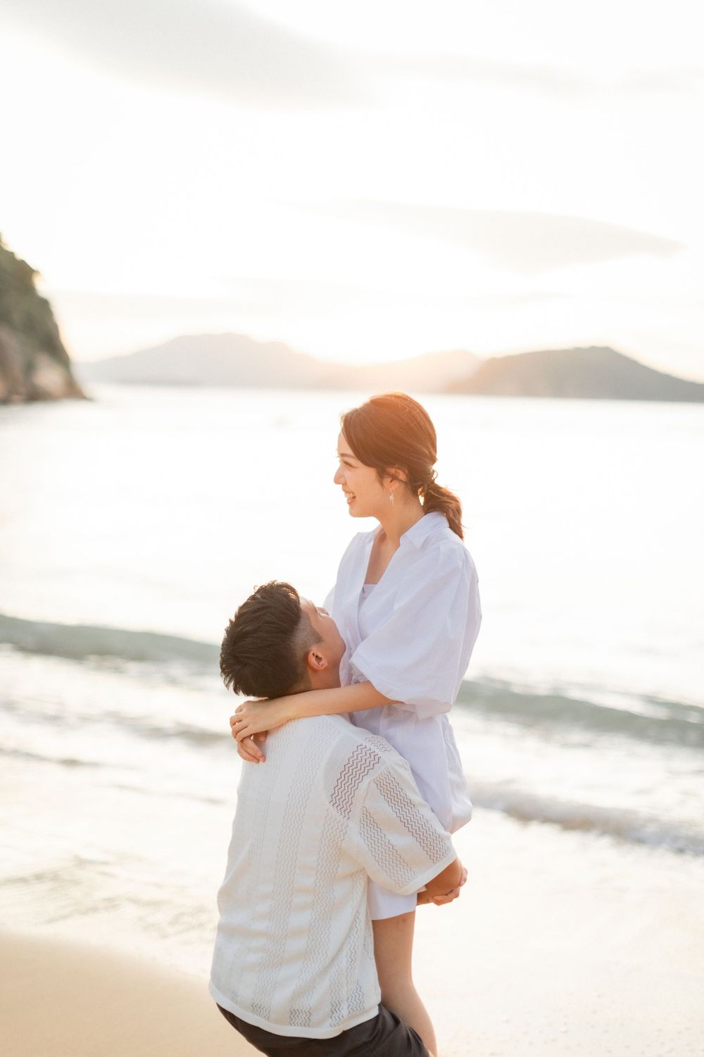Man lifting woman on a beach at sunset, both smiling and dressed in white casual outfits.