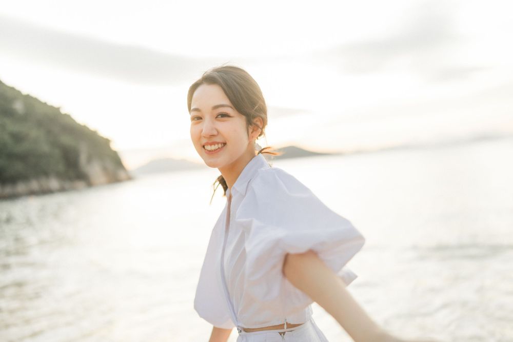 Smiling Asian woman in a white dress posing on a beach at sunset with ocean and hills behind her.