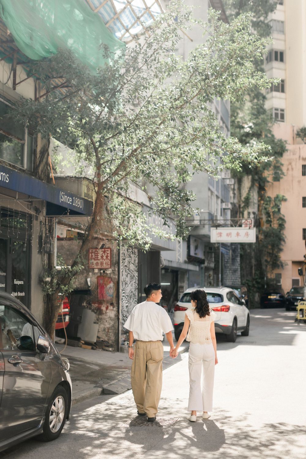 Asian couple holding hands while walking along a sunny urban street lined with trees.