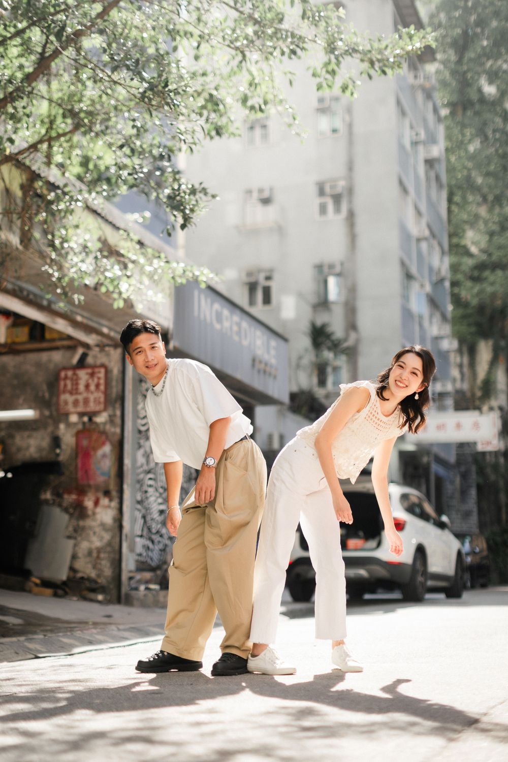 Playful Asian couple laughing and posing on a sunlit urban street with buildings behind.