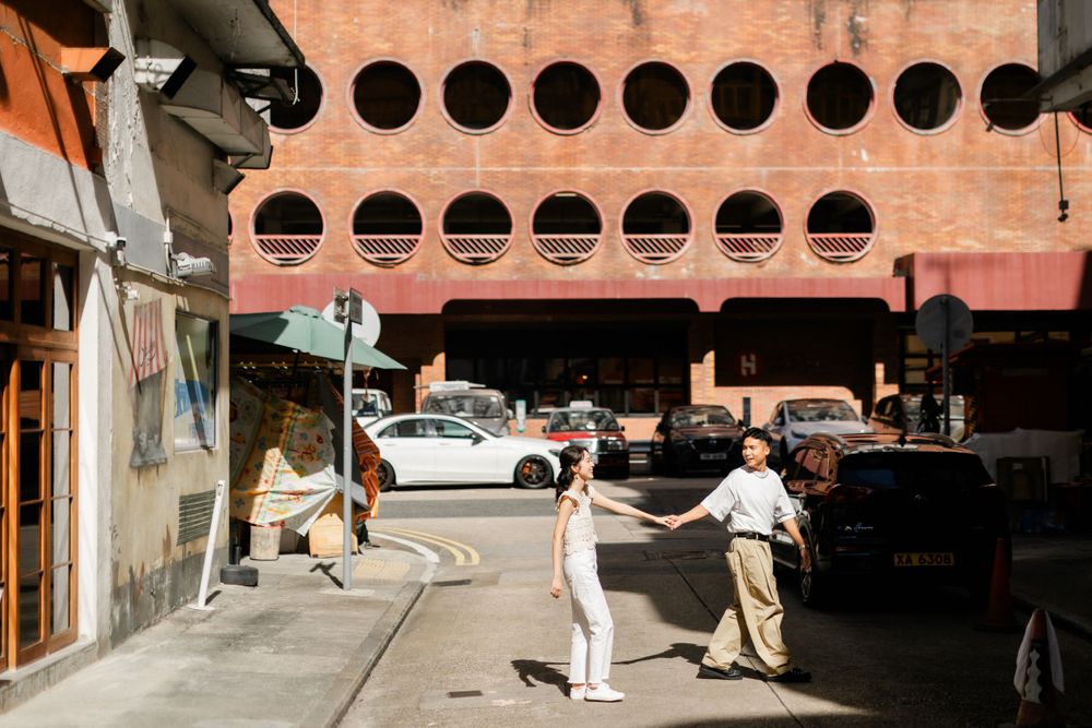 Asian couple holding hands while crossing a street near a distinctive circular-windowed building.