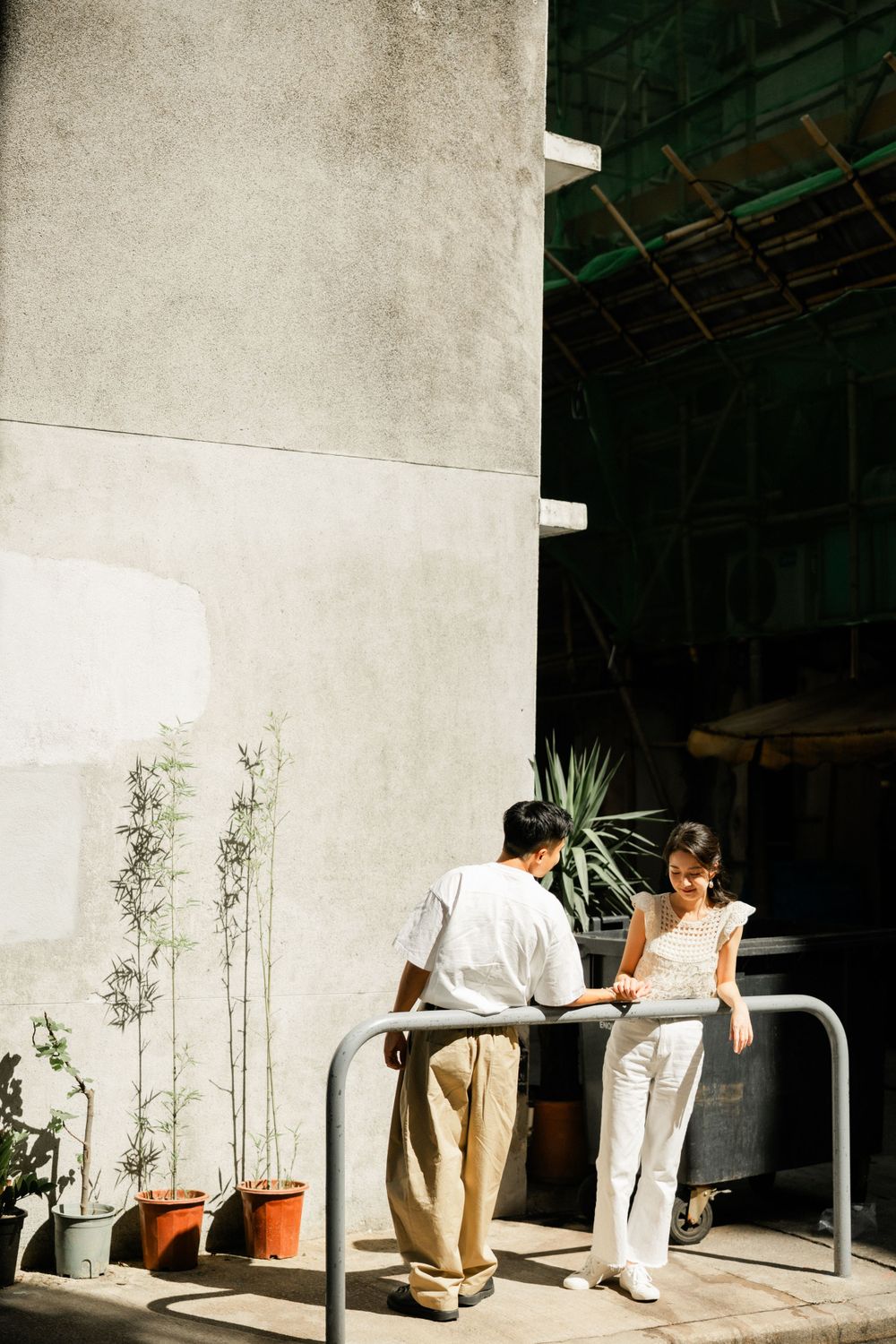 Asian couple leaning on a metal railing outside a sunlit building surrounded by potted bamboo plants.