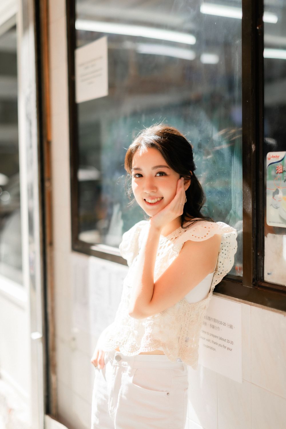 Smiling young Asian woman in white lace top posing beside a window in warm sunlight.