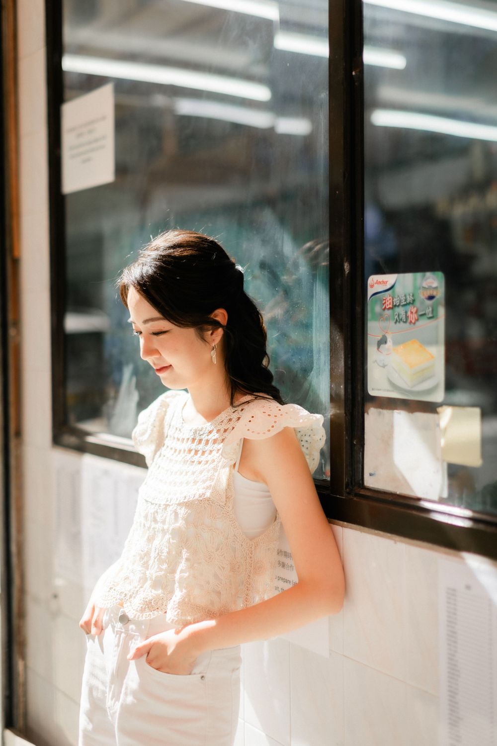 Elegant Asian woman in white crochet top standing thoughtfully beside a shop window outdoors.