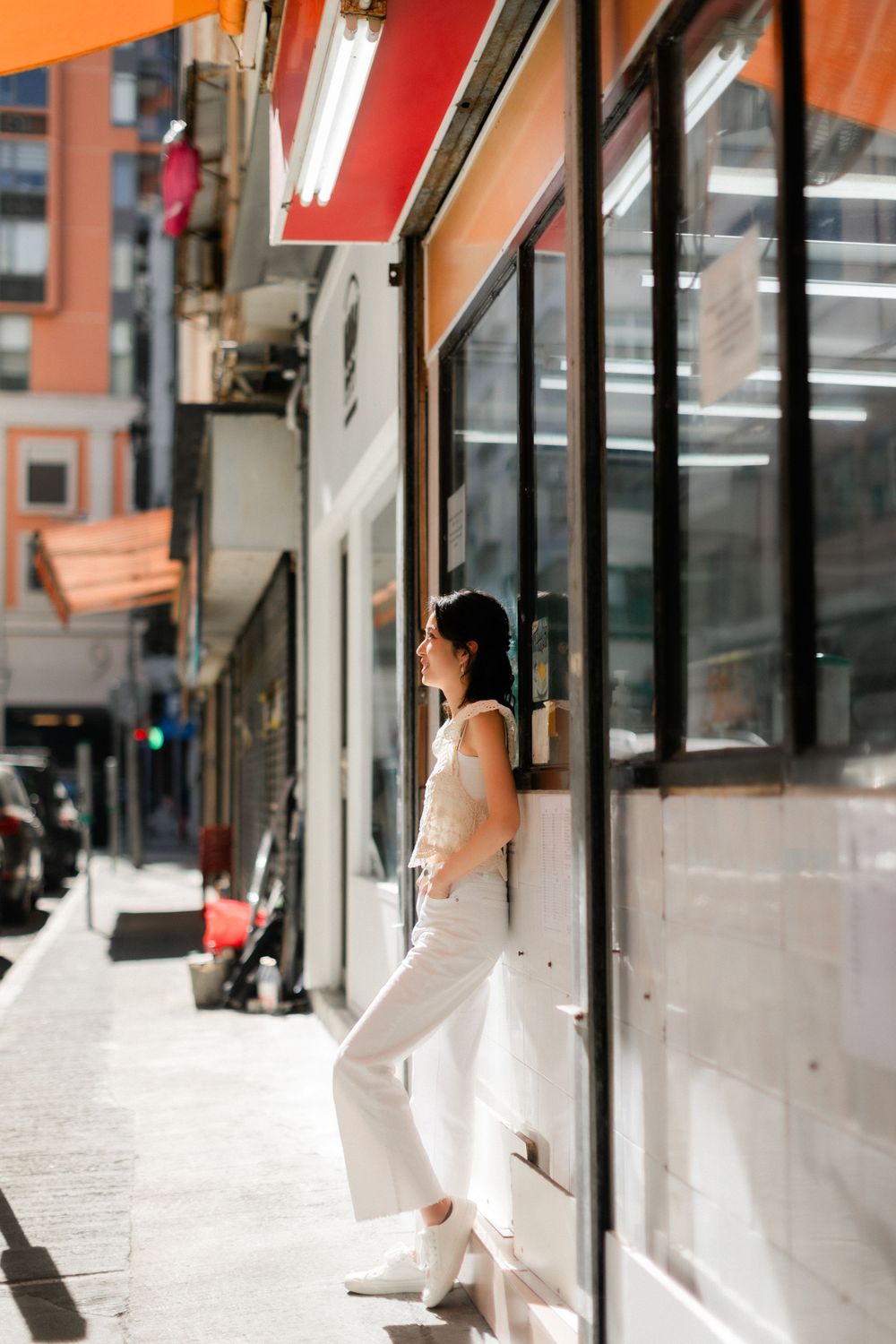 Stylish Asian woman in white outfit leaning against a storefront on a sunny city street.
