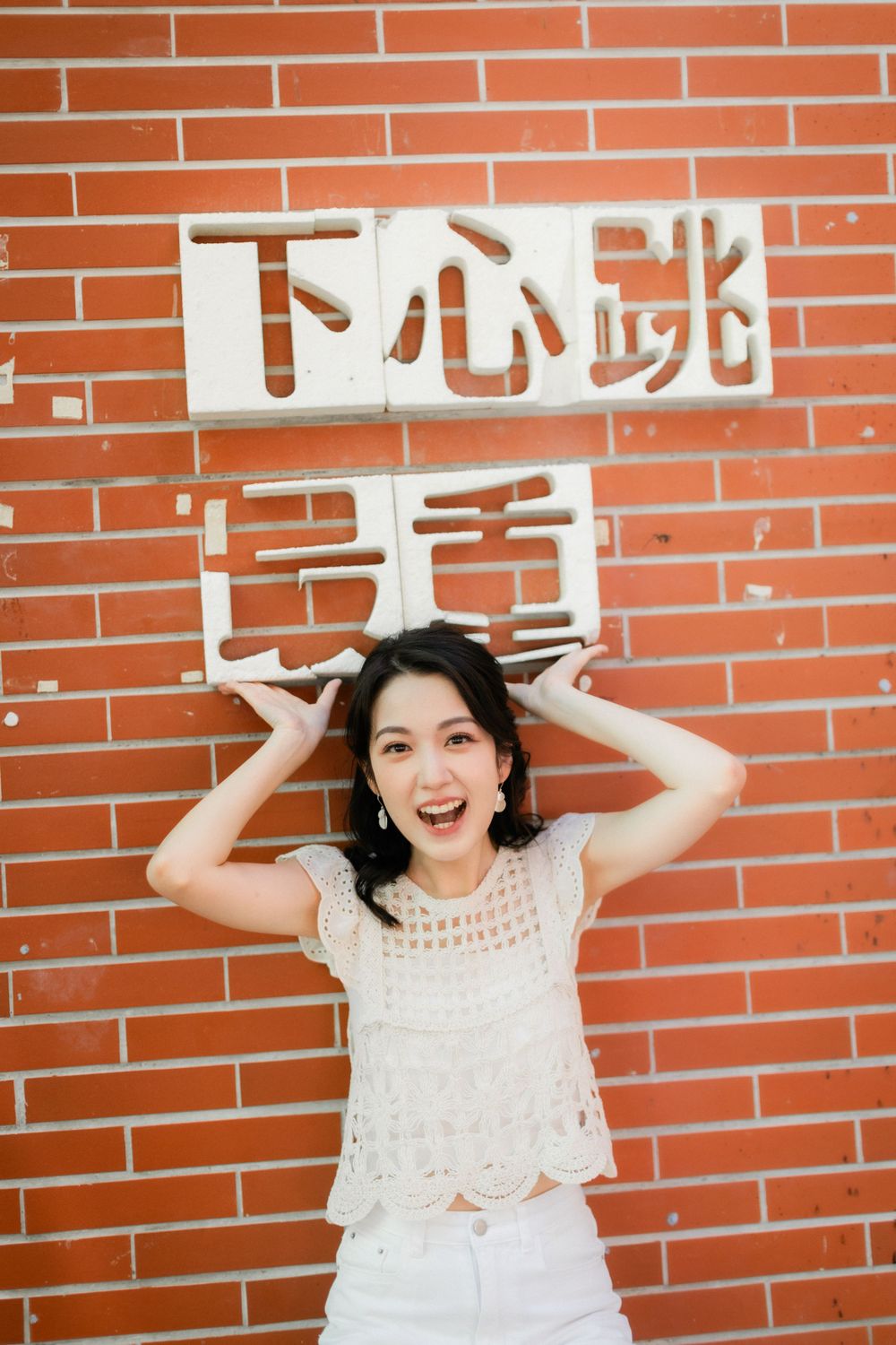 Joyful young Asian woman in white lace top posing with Chinese characters sign on red brick wall.