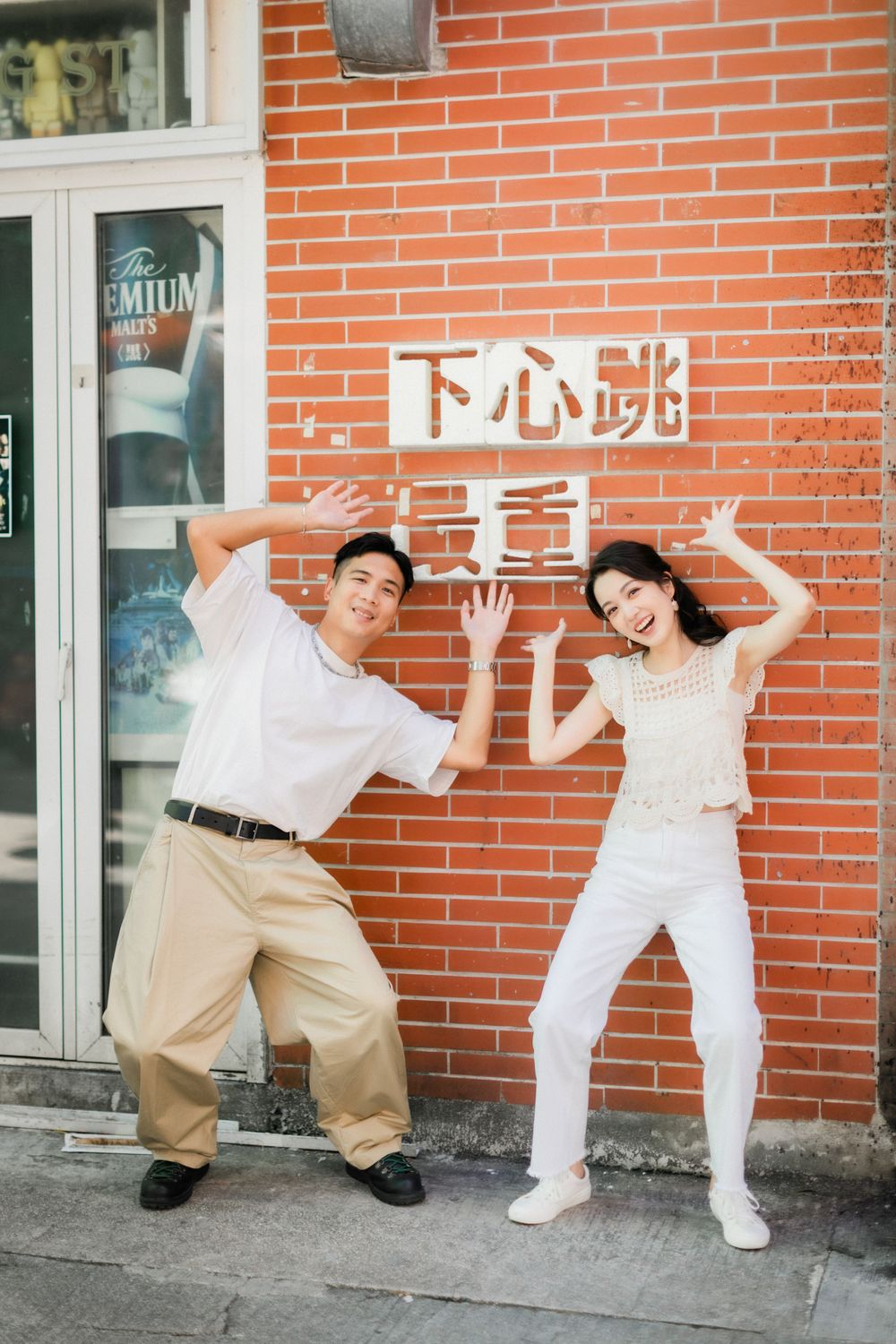 Happy Asian couple posing playfully in front of red brick wall with Chinese characters sign.