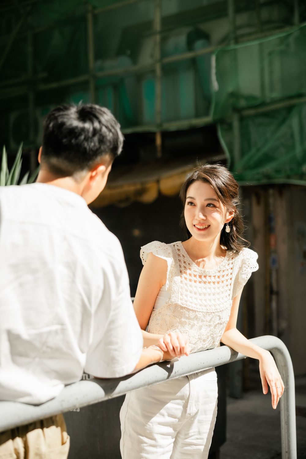 Smiling Asian woman in a white lace top leaning on a metal railing while talking to a man outdoors.