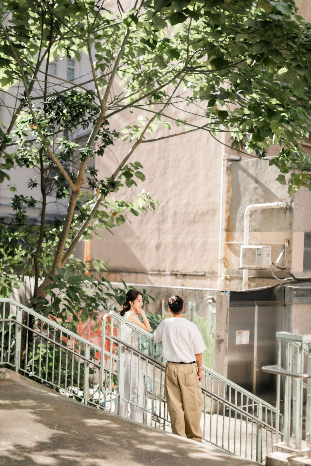 Asian couple standing on outdoor staircase surrounded by lush green trees in urban setting.
