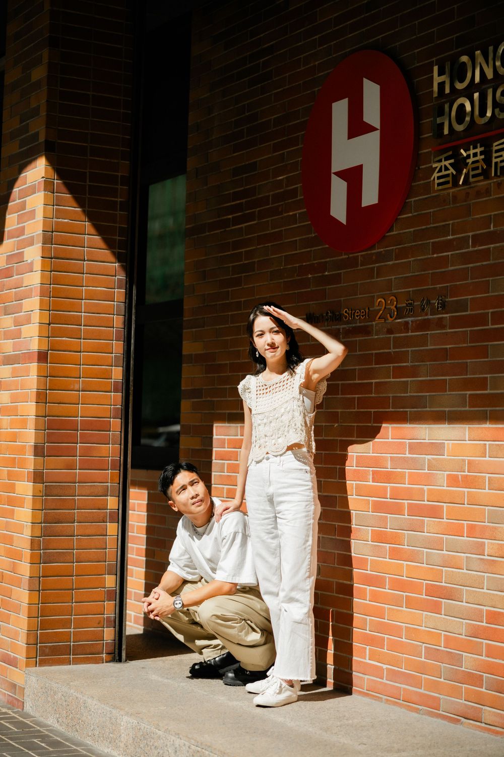 Asian couple posing in front of a red brick Hong Kong House building entrance in casual white outfits.