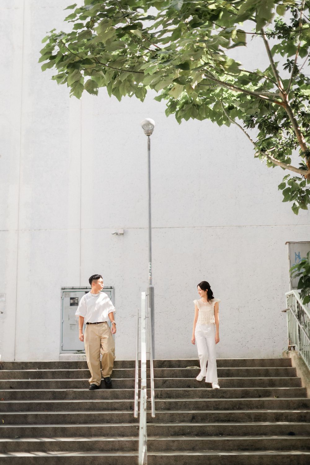 Asian couple walking down outdoor stairs with green trees and white wall in background.
