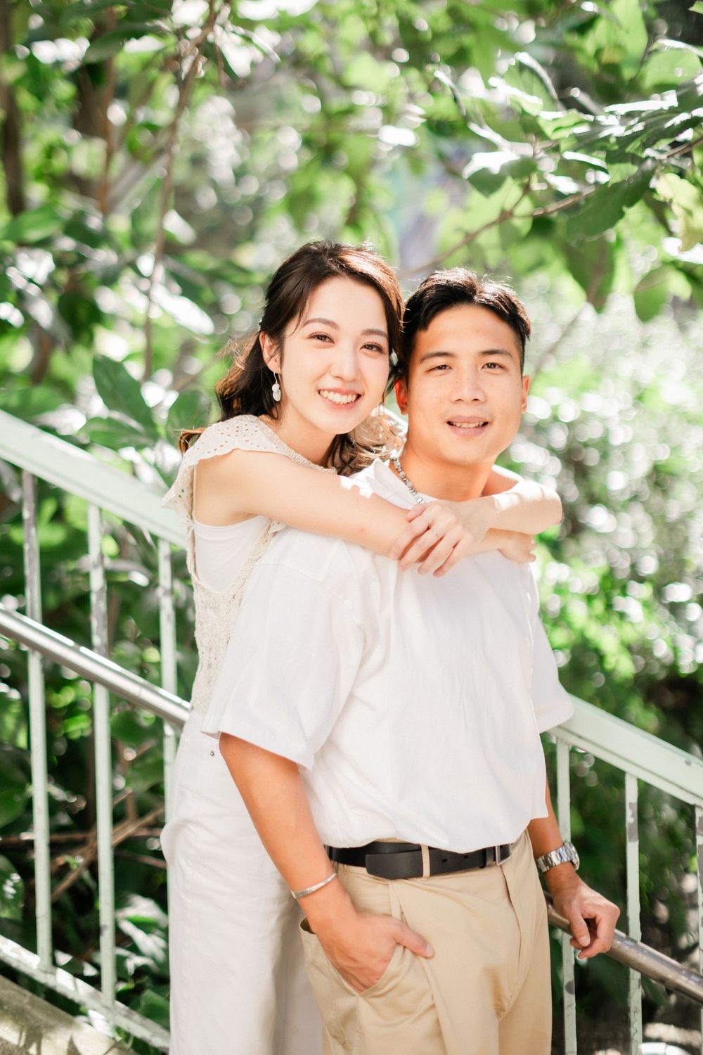 Smiling Asian couple embracing on staircase surrounded by green foliage in natural light.
