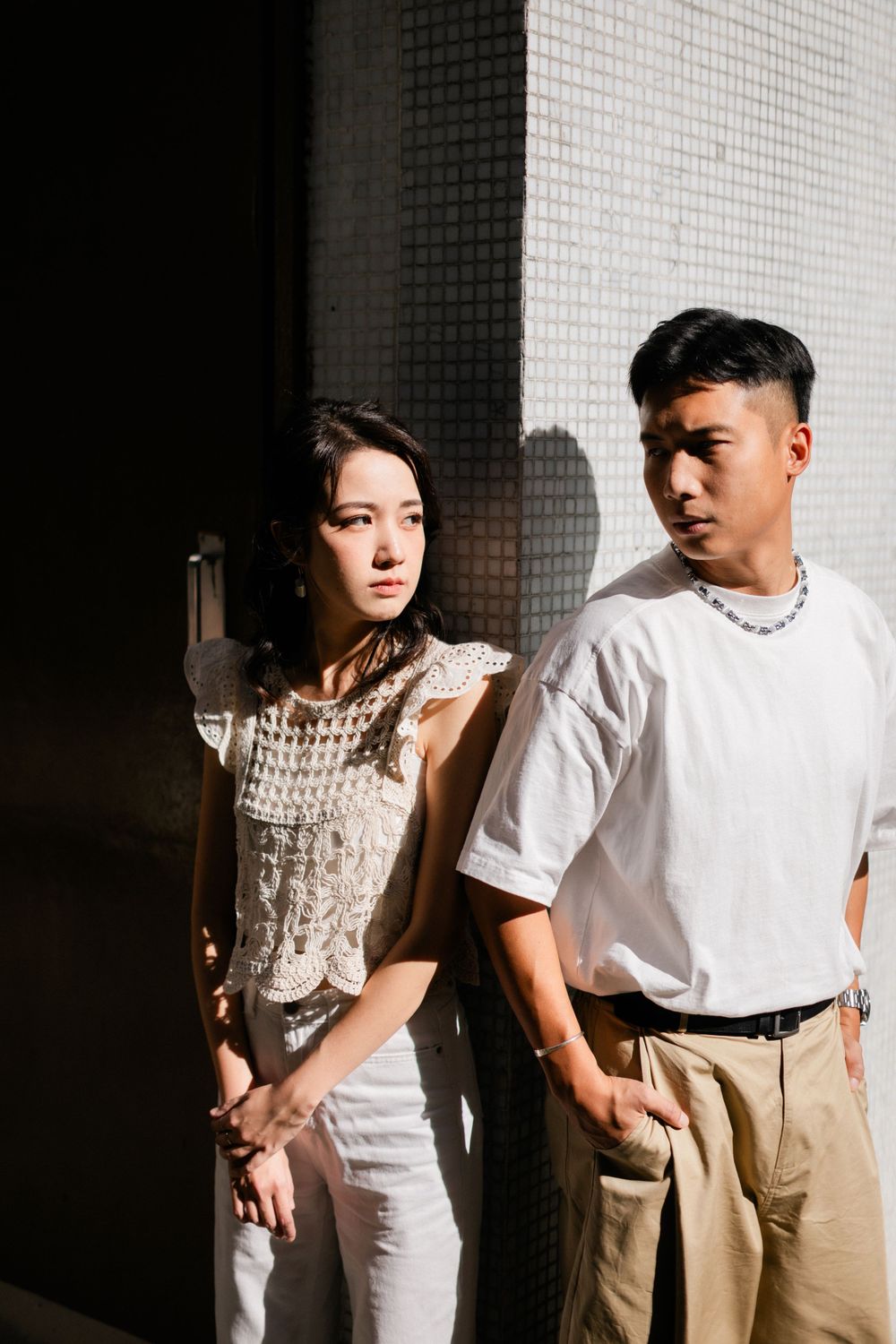 Young Asian couple posing in dramatic sunlight and shadow against white tiled wall.