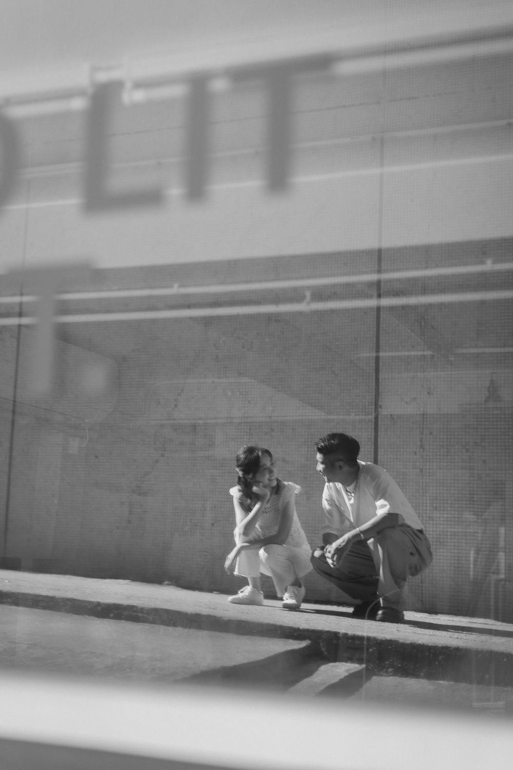 Black and white photo of a couple crouching together outside a building with reflective glass windows.