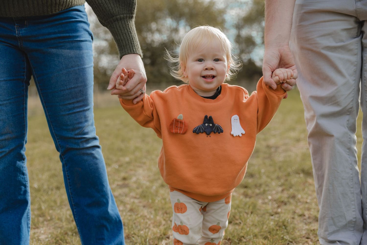 A toddler in orange sweater walking while holding hands in a grassy field.  Photographer. Hull. East Yorkshire. Photography