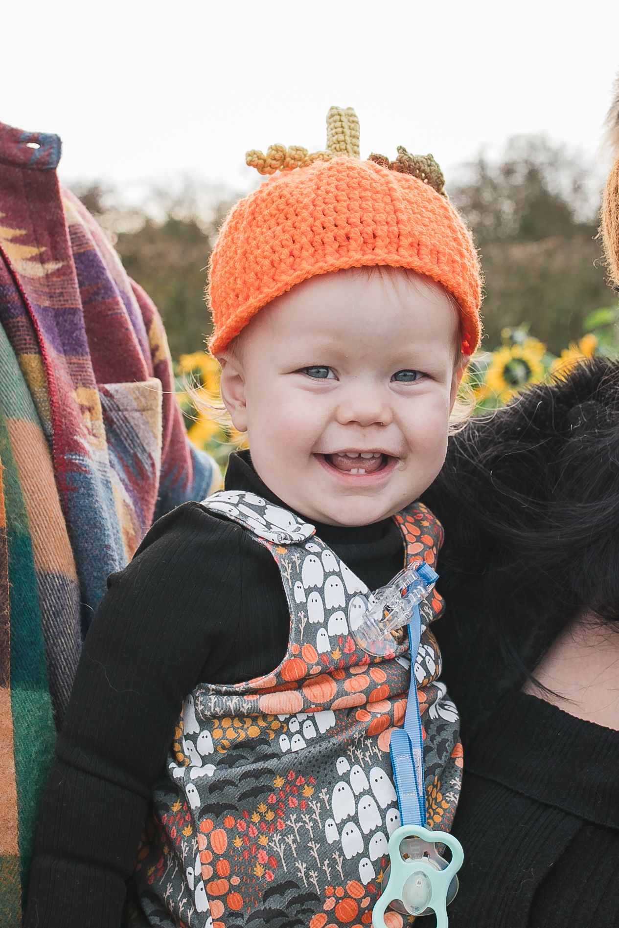 Baby wearing an orange knit hat and patterned outfit smiles while being held in a pumpkin patch. Photographer. Hull. East Yorkshire. Photo
