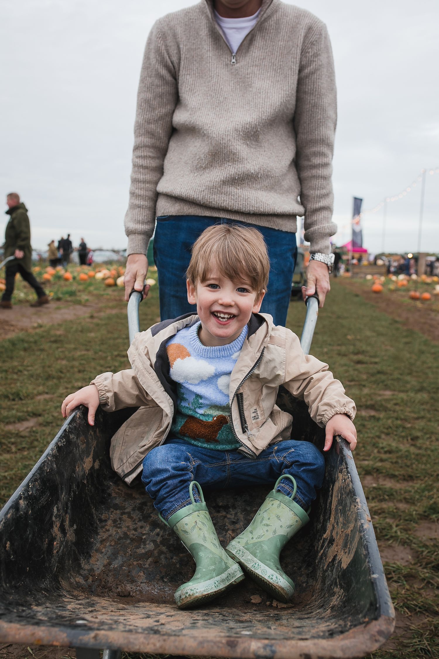 Child wearing green boots sits in a muddy wheelbarrow while wearing casual outdoor clothing. Photographer. Hull. East Yorkshire. Photography