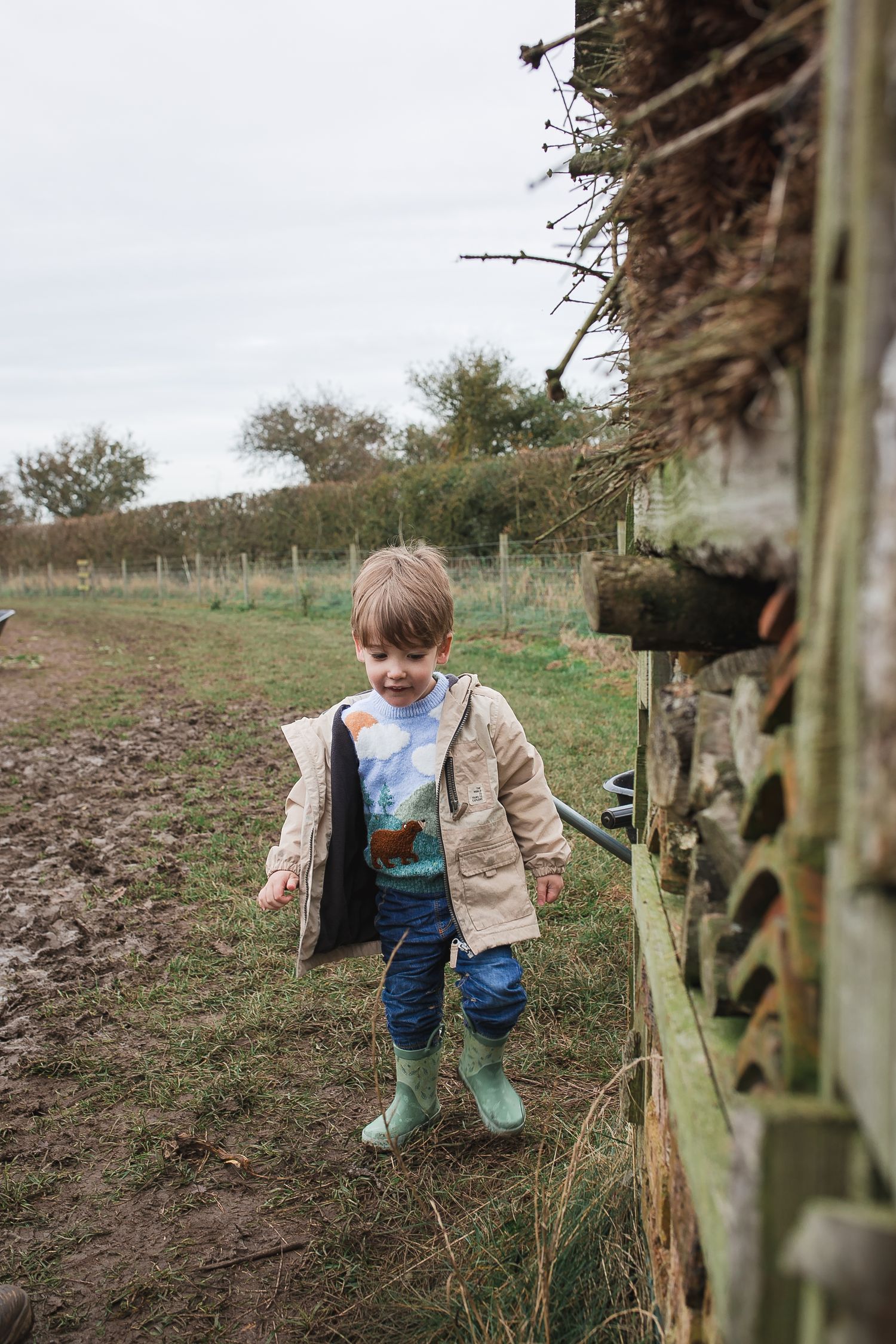 Child in green boots and tan jacket plays near wooden fence posts in muddy field. Photographer. Hull. East Yorkshire. Photography