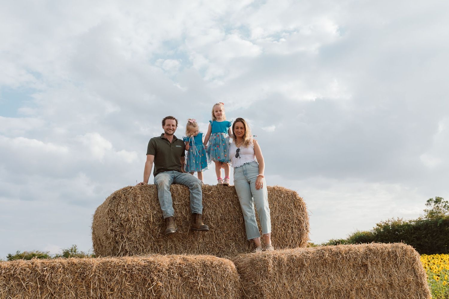 Family poses atop stacked hay bales with sunflower fields in background. Photographer. Hull. East Yorkshire. Photography
