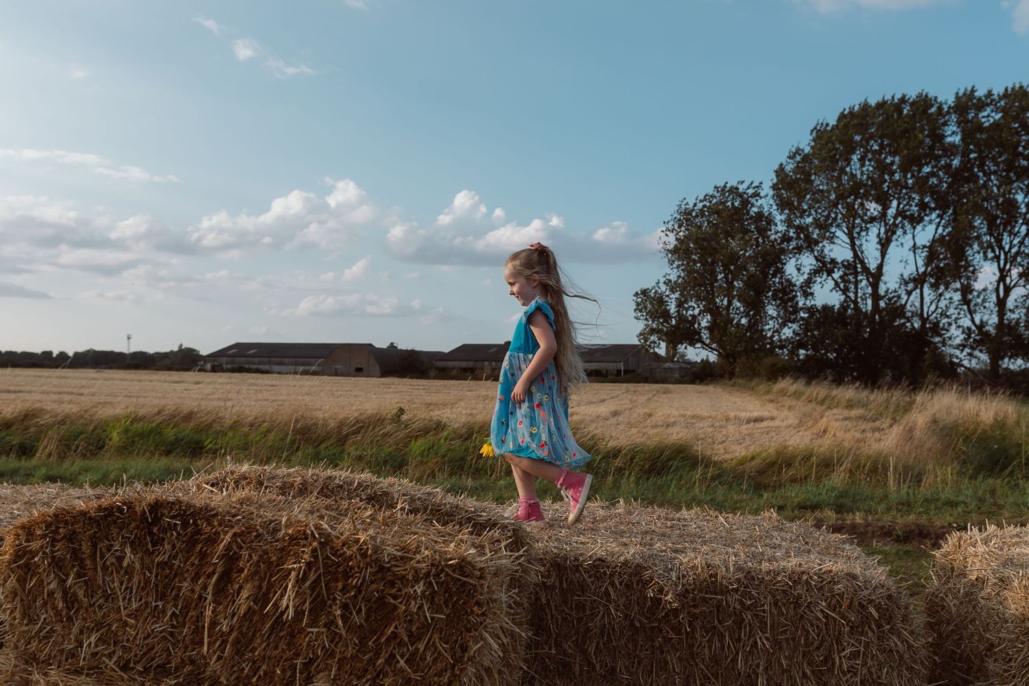 Child in blue dress stands on hay bale against cloudy countryside sky. Photographer. Hull. East Yorkshire. Photography