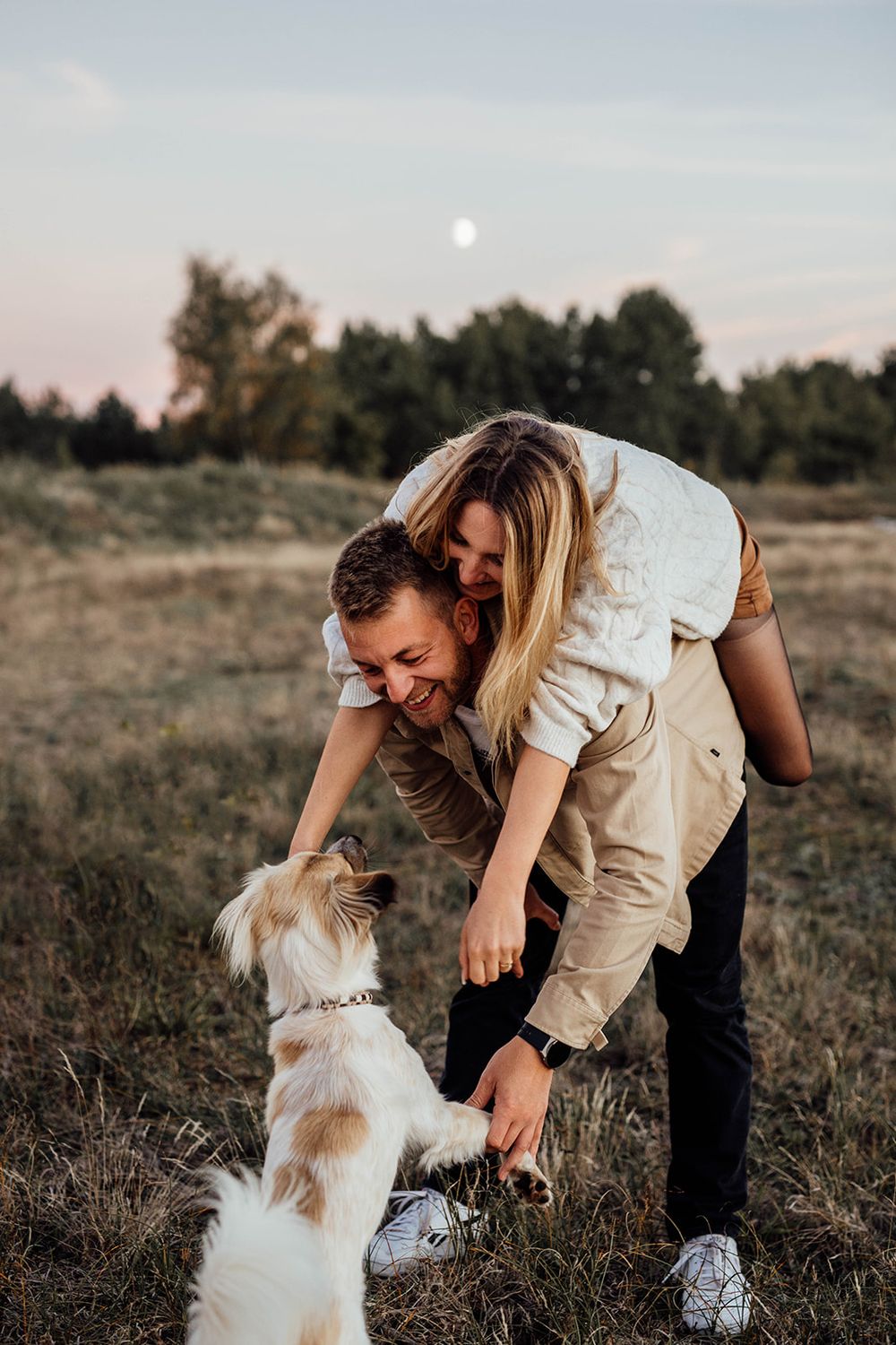 A couple plays with their small white and tan dog in an open field at dusk with the moon visible in the sky.
