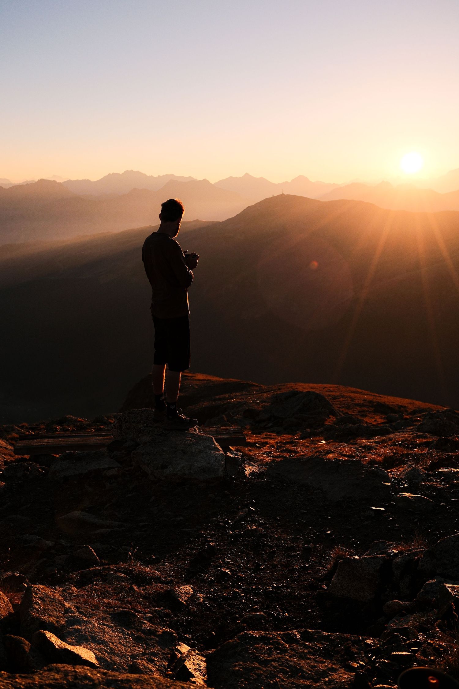 Silhouetted hiker stands on mountain summit against dramatic golden sunset with mountain ranges in background.