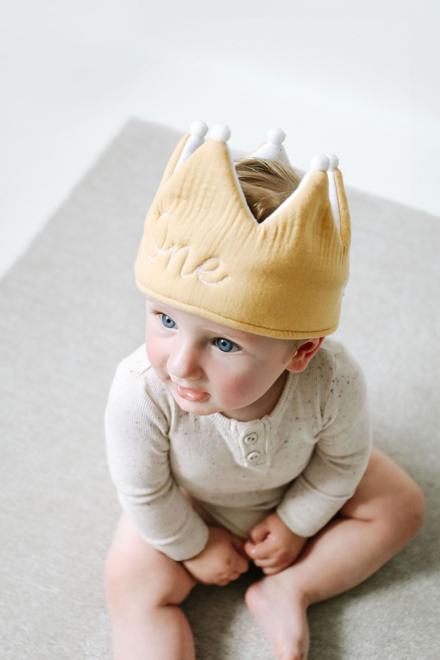 A small child wearing a pale yellow fabric "one" crown and light coloured clothing sits on a grey surface during birthday shoot in Nottingha