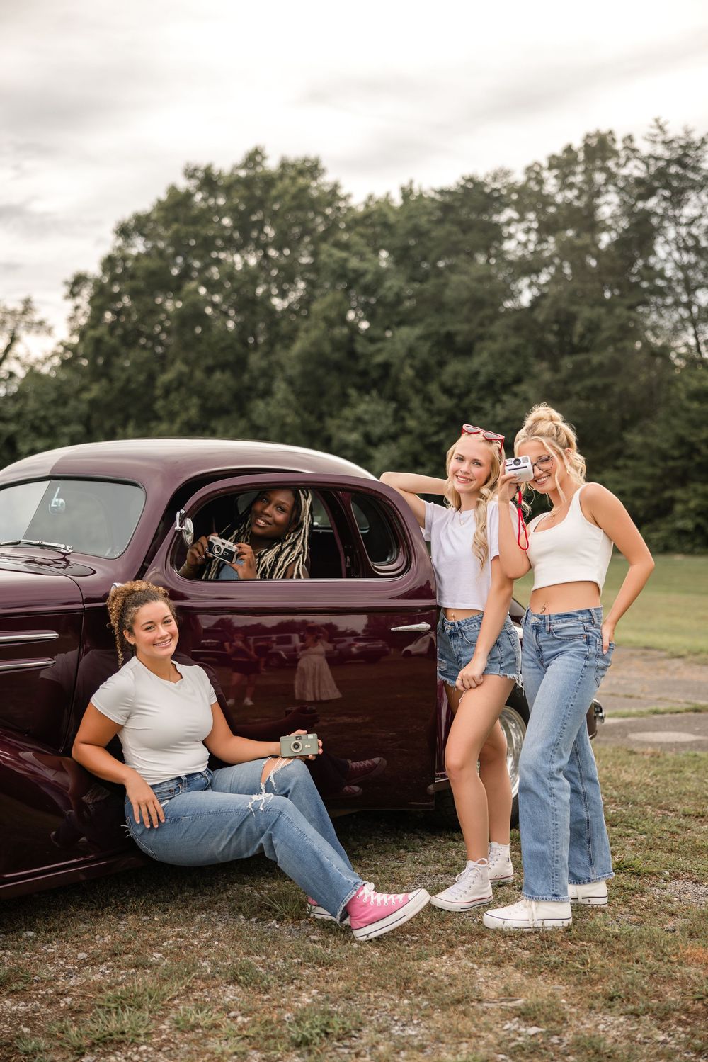Group of friends gather around a vintage maroon car at a rural outdoor location with trees in the background.