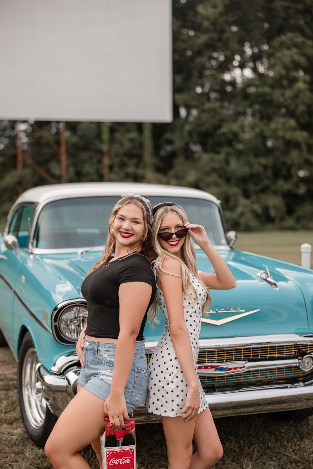 Two friends pose with a classic turquoise Chevrolet car while holding a Coca-Cola bottle.