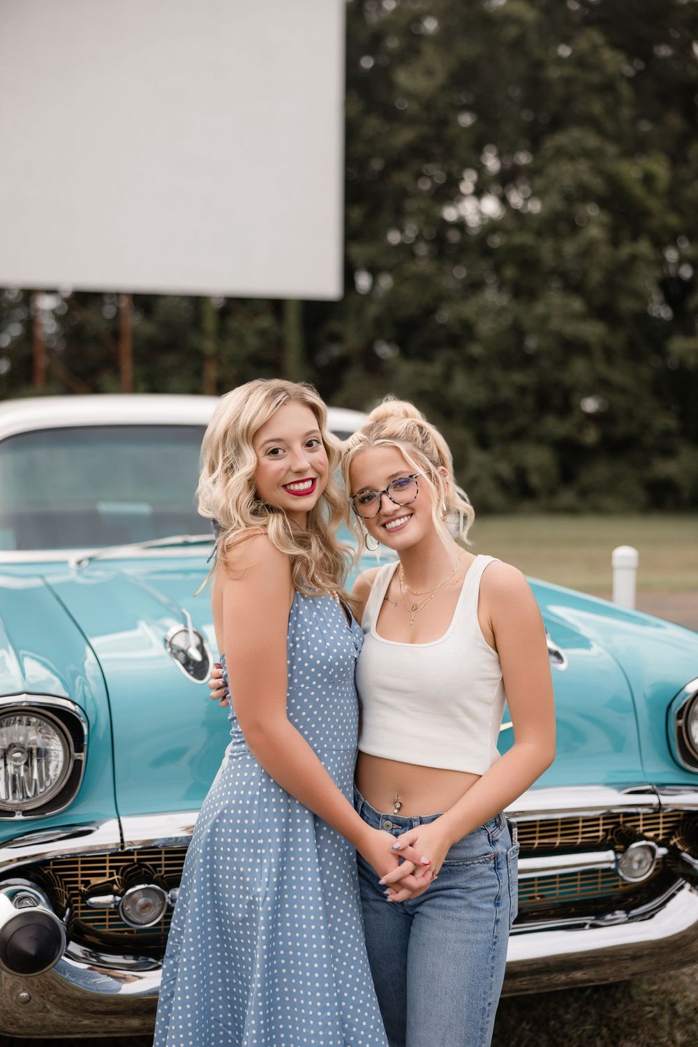Two friends pose together next to a vintage turquoise classic car in a casual outdoor photoshoot.