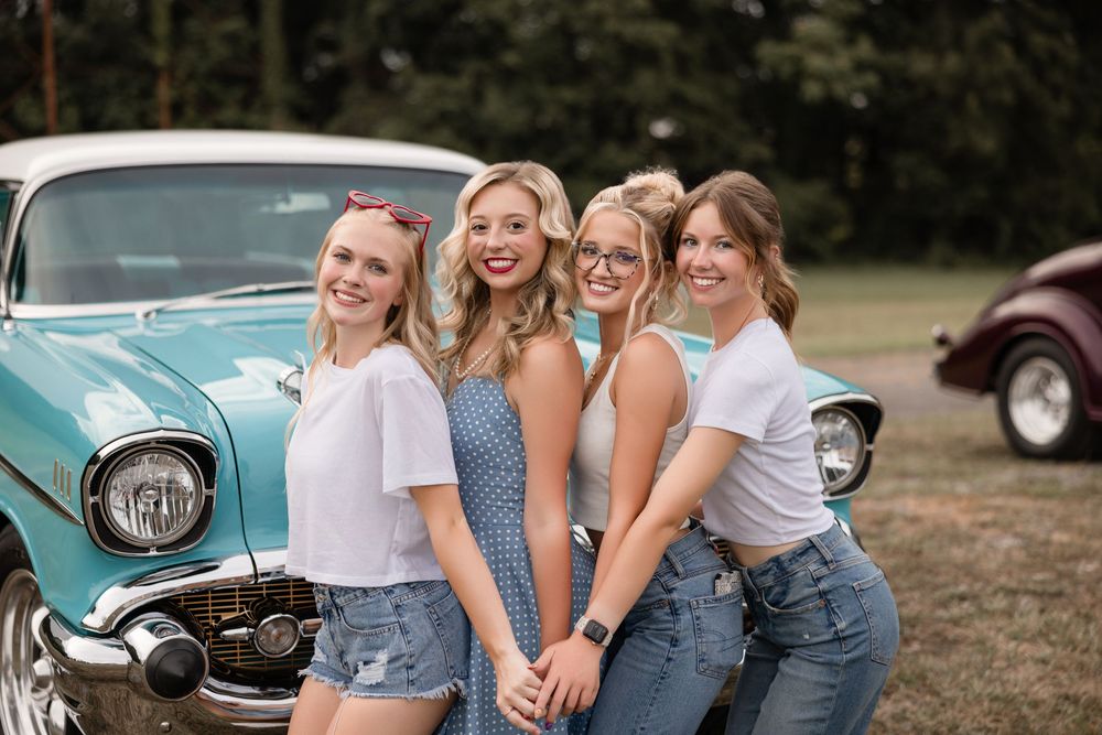 Four friends pose together in front of a classic turquoise vintage car on a sunny summer day.