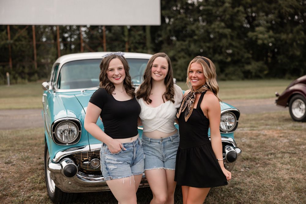 Three friends stand together smiling in front of a vintage turquoise car during a summer photo shoot.