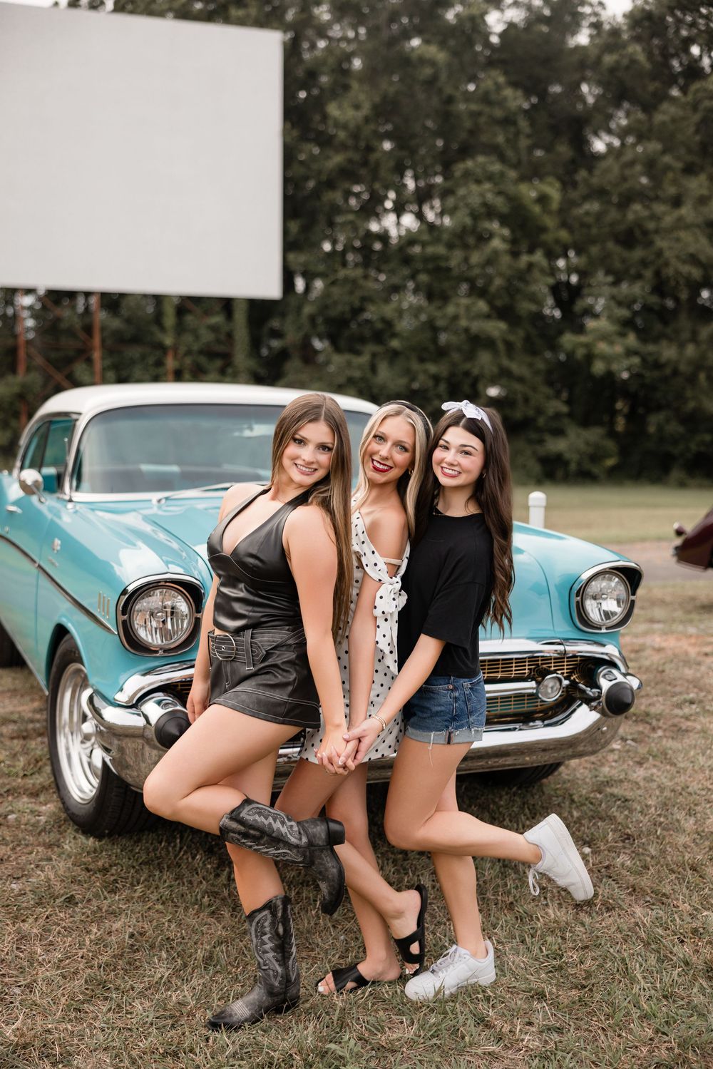 Three friends pose together in casual summer outfits next to a classic turquoise Chevrolet car at an outdoor location.