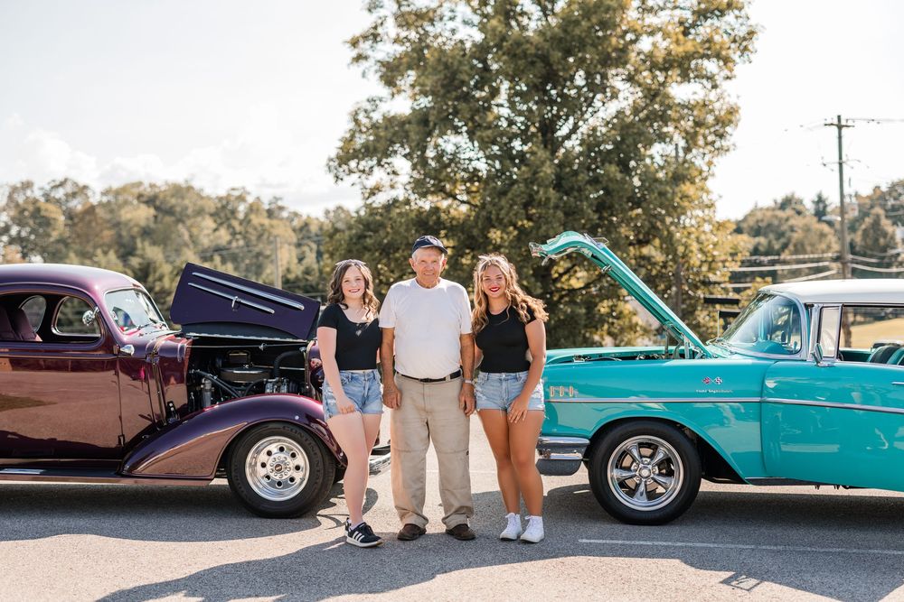 Three people pose between a vintage brown hotrod and turquoise classic car at a car show event.