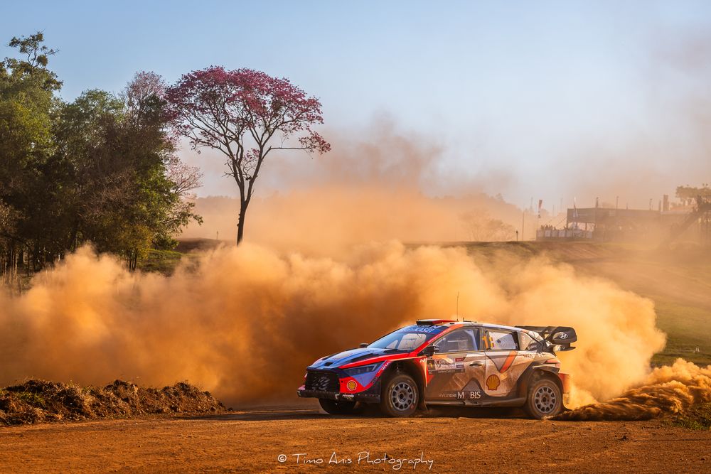A Hyundai rally car kicks up orange dust while navigating a challenging dirt track turn.