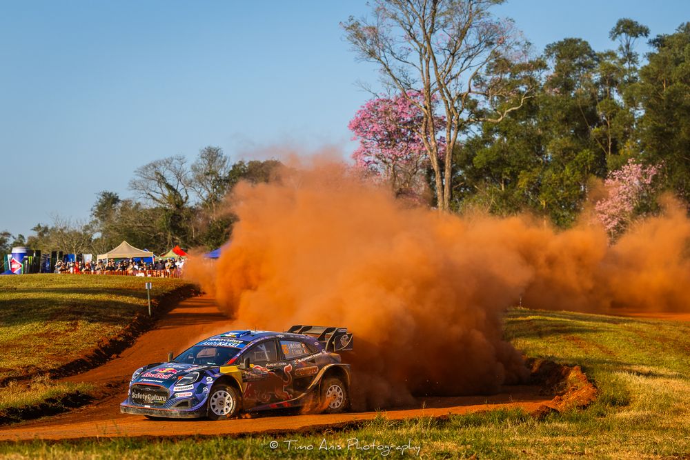 Rally car kicks up clouds of orange dust while racing through a dirt track during a high-speed motorsport competition.