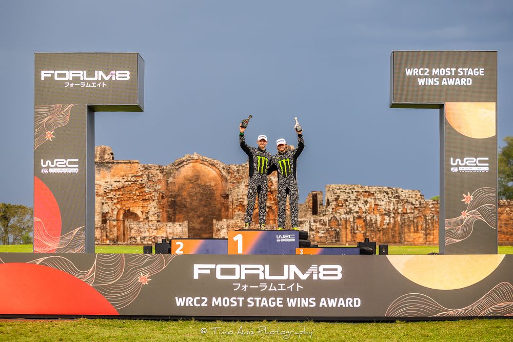 WRC2 stage win award ceremony showing competitors standing on a podium with graphics and banners.