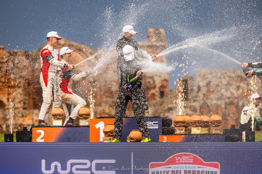 Champagne celebration on motorsport podium with winners spraying bottles in victory celebration.