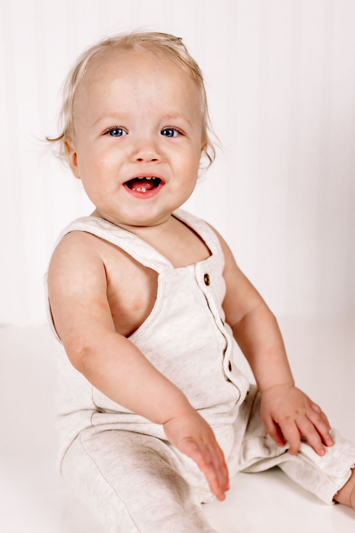 Happy baby wearing white sleeveless romper sitting against light background while laughing and smiling.