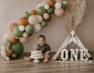 A boho-themed first birthday party setup with earth-toned balloon garland, teepee tent, and white cake display.