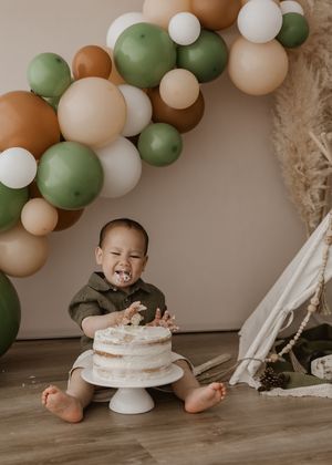 Baby in white outfit sits on floor with green, peach and white balloon arch for cake smash photo session.