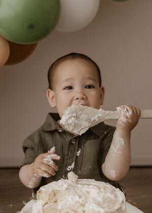 Small child in green shirt enjoys smashing and playing with white frosted birthday cake at party with balloons.